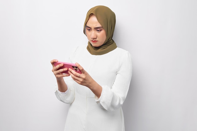 Ritratto di donna musulmana asiatica infastidita che indossa l'hijab che gioca con il telefono cellulare isolato su sfondo bianco ritratto in studio Persone concetto di stile di vita religioso