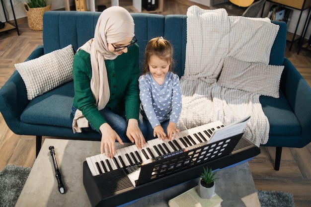 Ritratto di donna musulmana in hijab e la sua piccola figlia carina che impara a suonare il pianoforte a casa