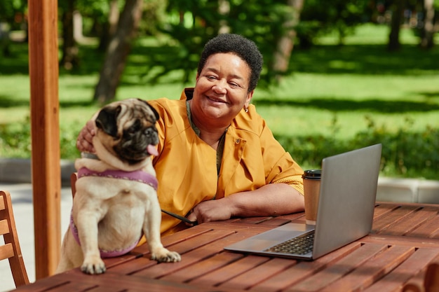 Ritratto di donna nera sorridente che usa il laptop in un caffè all'aperto con una scena di cane carlino carino illuminata dalla luce del sole