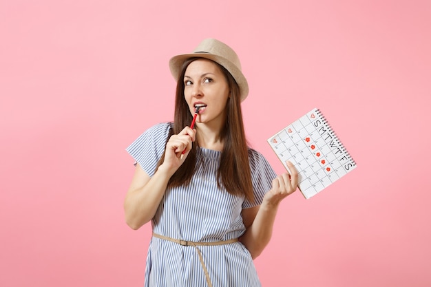 Ritratto di donna pensierosa in abito blu, cappello con matita rossa, calendario dei periodi femminili per controllare i giorni delle mestruazioni isolati su sfondo rosa. Sanità medica, concetto ginecologico. Copia spazio