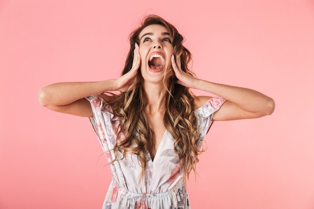 Ritratto di donna positiva con capelli lunghi che indossa abito in piedi e urlando, isolato in rosa