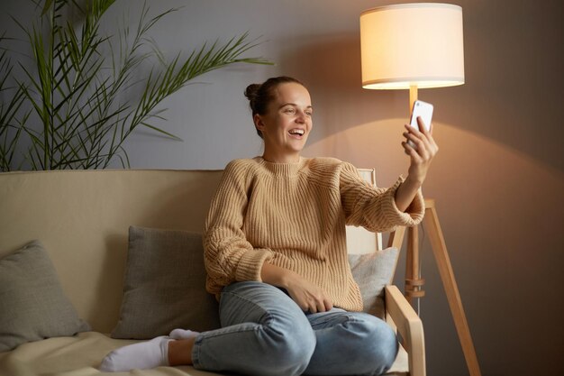 Ritratto di donna positiva estremamente felice che indossa un maglione beige e jeans seduti sulla tosse e prendendo selfie o facendo una videochiamata ridendo essendo di buon umore