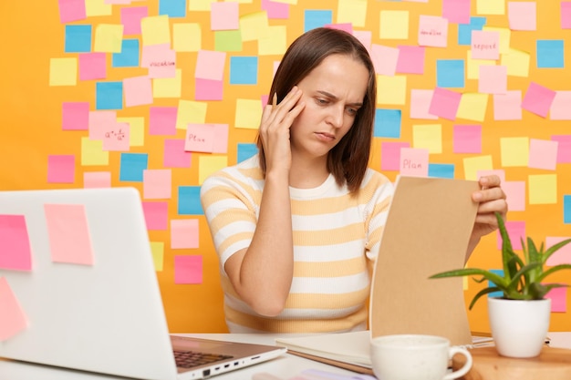 Ritratto di donna premurosa con i capelli castani che indossa una maglietta a righe seduta al tavolo in posa contro il muro giallo con piccole note adesive che lavorano in ufficio pensando durante la lettura di documenti