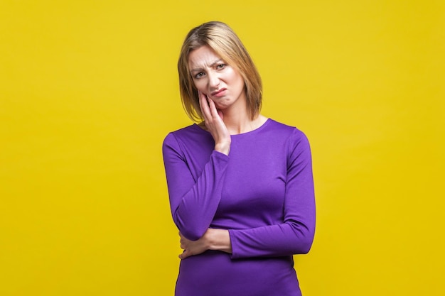 Ritratto di donna premurosa indifferente in elegante abito viola attillato che tiene la mano sulla guancia e sembra disinteressato e insoddisfatto girato in studio al coperto isolato su sfondo giallo