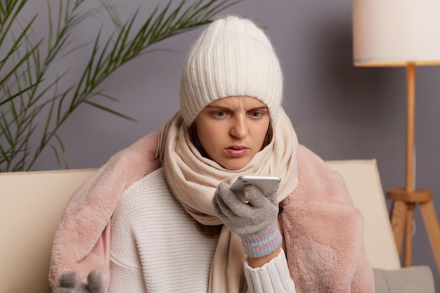 Ritratto di donna scioccata e spaventata seduta sul divano di casa che indossa un cappotto invernale cappello e guanti che tengono il telefono cellulare guardando il display con occhi grandi