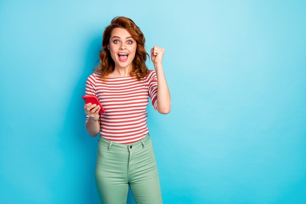Ritratto di donna scioccata felice utilizzare il cellulare vincere lotteria dei social media alza i pugni urlare sì indossare pullover elegante isolato su colore blu