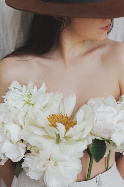 Ritratto di donna sensuale alla moda in cappello con fiori di peonia bianca sul corpo Fragranza floreale
