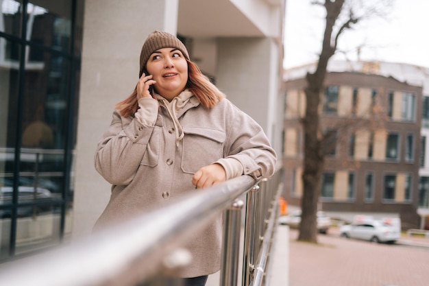 Ritratto di donna seria in sovrappeso con cappello caldo e giacca che parla al telefono cellulare in piedi vicino alla ringhiera in una strada cittadina in una nuvolosa giornata autunnale. Femmina congelata che ha chiamata sullo smartphone all'aperto.