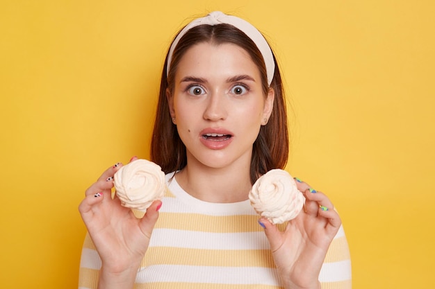 Ritratto di donna sorpresa scioccata che indossa fascia per capelli e maglietta a righe in piedi tenendo marshmallow e guardando la fotocamera con occhi grandi e bocca aperta isolata su sfondo giallo