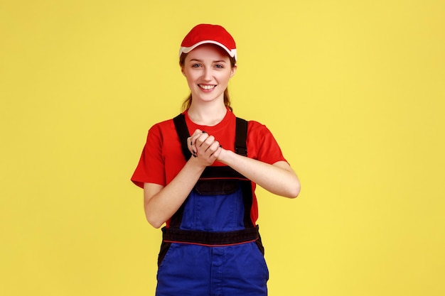 Ritratto di donna sorridente a portata di mano che preme le mani insieme sul petto godendo, calmandosi e confortando se stessa, indossando tute e berretto rosso. Studio indoor girato isolato su sfondo giallo.