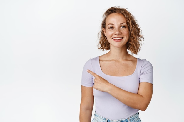 Ritratto di donna sorridente che mostra vendita, puntando il dito a sinistra, prodotto pubblicitario, in piedi in maglietta su sfondo bianco