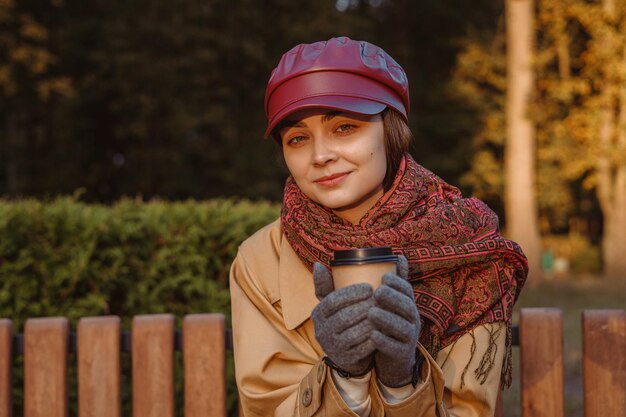 Ritratto di donna sorridente che scalda le mani con una tazza di caffè per andare e guardare la fotocamera nel parco autunnale