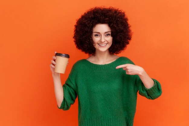 Ritratto di donna sorridente con acconciatura afro che indossa un maglione verde in stile casual in piedi che punta a una tazza usa e getta con caffè da asporto. Studio indoor girato isolato su sfondo arancione.