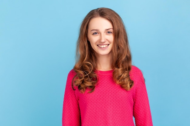 Ritratto di donna sorridente con capelli ondulati che indossa un pullover rosa che esprime emozioni positive e felicità, guardando la fotocamera, essendo di buon umore. Studio indoor girato isolato su sfondo blu.