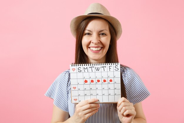 Ritratto di donna sorridente in abito blu, cappello che tiene il calendario dei periodi per controllare i giorni delle mestruazioni isolati su sfondo rosa di tendenza brillante. Sanità medica, concetto ginecologico. Copia spazio.