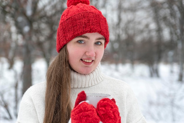 Ritratto di donna sorridente in cappello di lana rosso e guanti che tengono una tazza di bevanda calda all'esterno