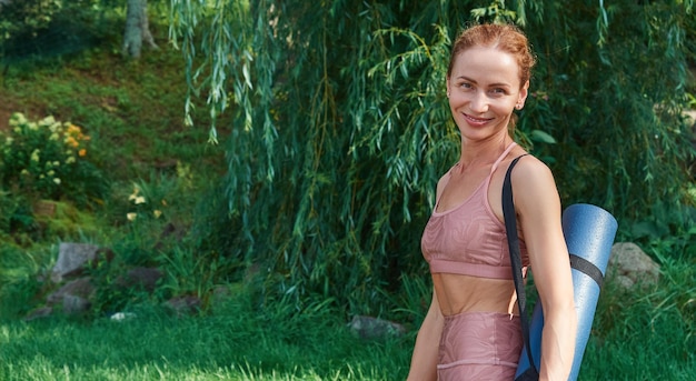 Ritratto di donna sportiva che tiene tappetino da yoga guardando la fotocamera sorridente mentre si prepara per l'allenamento all'aperto il giorno d'estate