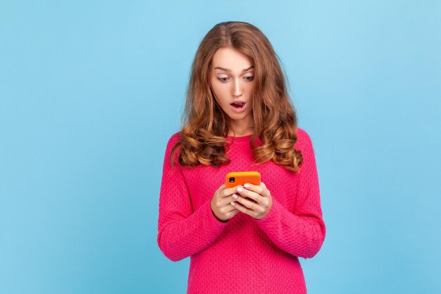 Ritratto di donna stupita che indossa un pullover rosa, guardando il cellulare con espressione sorpresa, leggendo notizie scioccanti utilizzando il telefono cellulare. Studio indoor girato isolato su sfondo blu.