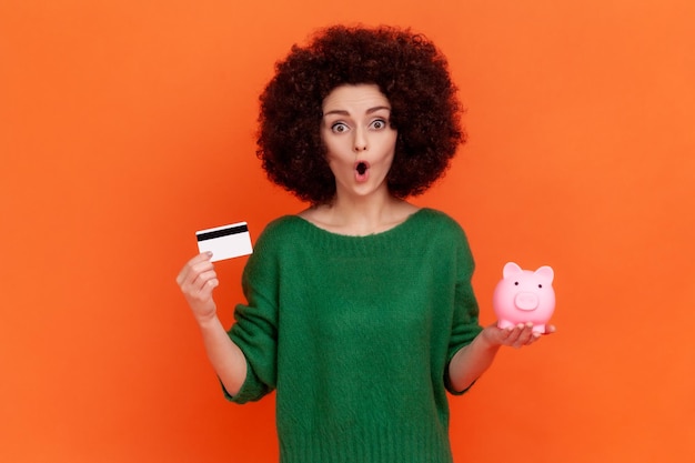 Ritratto di donna stupita con acconciatura afro in maglione verde stile casual in piedi con la bocca aperta, in possesso di carta di credito e salvadanaio, bancario. Studio indoor girato isolato su sfondo arancione.