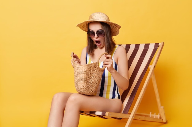 Ritratto di donna stupita e scioccata in costume da bagno cappello di paglia e occhiali da sole seduti sulla sedia a sdraio guardando all'interno della borsa con la bocca aperta dimenticato la protezione solare