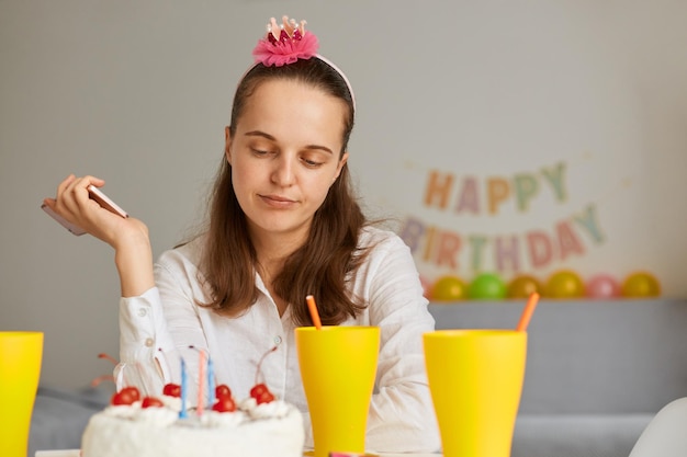 Ritratto di donna triste depressa che indossa abiti bianchi seduti a tavola con torta e bevanda, guardando il suo dessert di compleanno con espressione facciale sconvolta, festeggiando il compleanno da solo.