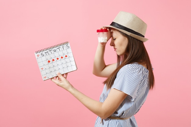 Ritratto di donna triste in abito blu che tiene una bottiglia bianca con pillole, calendario dei periodi femminili, controllando i giorni delle mestruazioni isolati sullo sfondo. Sanità medica, concetto ginecologico. Copia spazio.