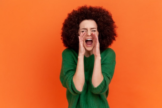 Ritratto di donna urlante arrabbiata con acconciatura afro che indossa un maglione verde in stile casual tenendo le mani vicino alla bocca, urlando ad alta voce. Studio indoor girato isolato su sfondo arancione.