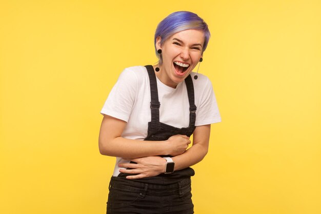 Ritratto di donna vivace e gioiosa hipster con capelli corti viola in tuta di jeans che tiene le braccia sulla pancia e ride ad alta voce divertito da scherzo divertente aneddoto isolato su sfondo giallo girato in studio