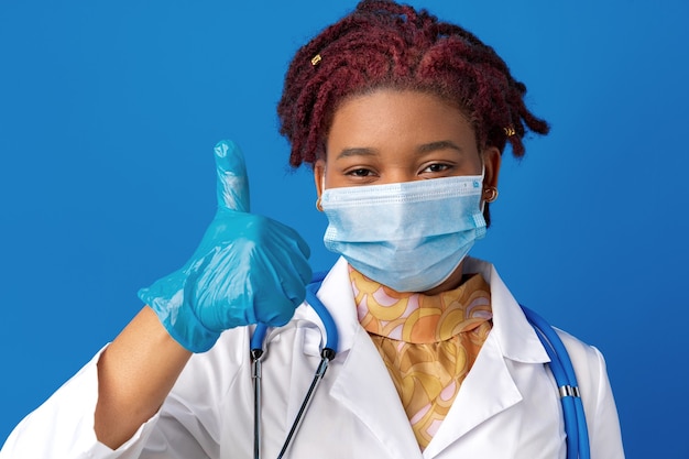 Ritratto di dottoressa africana in camice da laboratorio con maschera facciale e stetoscopio su sfondo blu