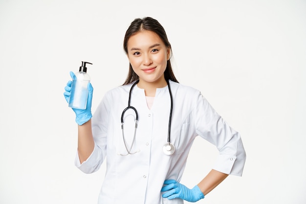 Ritratto di dottoressa asiatica, medico che raccomanda disinfettante per le mani per la prevenzione del covid-19, tenendo la bottiglia con guanti di gomma, sfondo bianco.