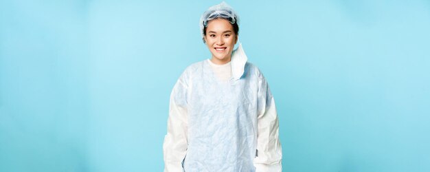 Ritratto di dottoressa o infermiera asiatica sorridente che indossa dispositivi di protezione individuale e che sembra felice in piedi su sfondo blu