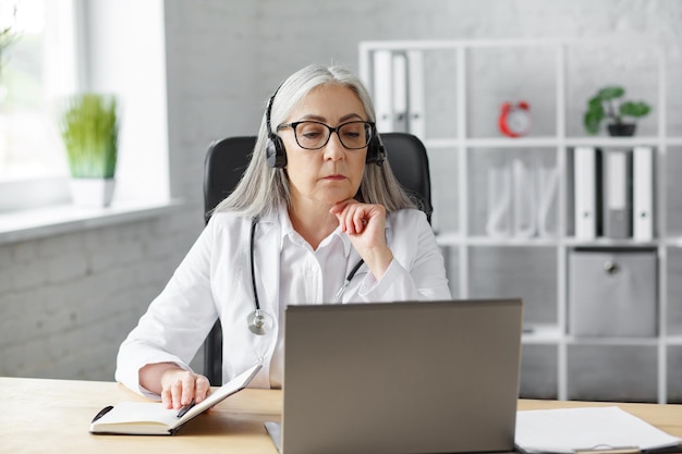 Ritratto di dottoressa senior dai capelli grigi nel suo ufficio che utilizza il laptop per la chat video con un paziente