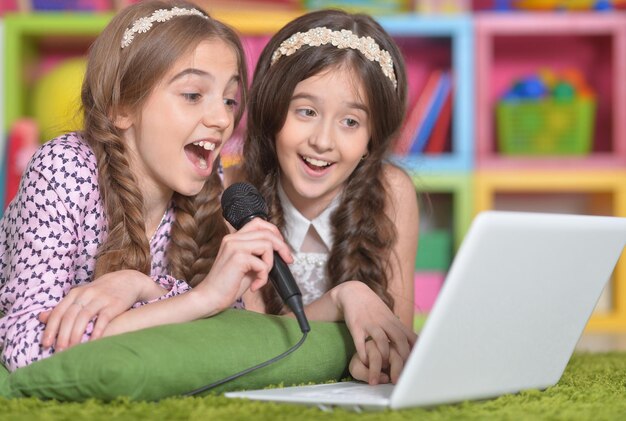 Ritratto di due bambine carine che cantano al karaoke con il computer portatile mentre sono sdraiate sul pavimento