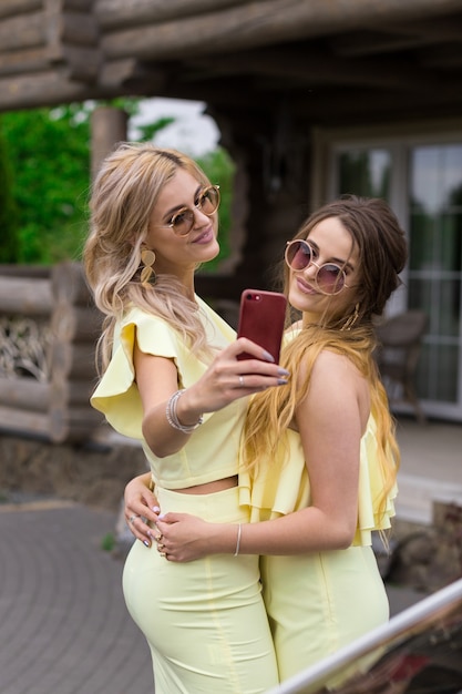 Ritratto di due damigelle d'onore piuttosto giovani in abiti gialli vicino a una casa in legno alla moda. Persone concetto di stile di vita. Facendo selfie sul cellulare sorridendo e inviando un bacio d'aria