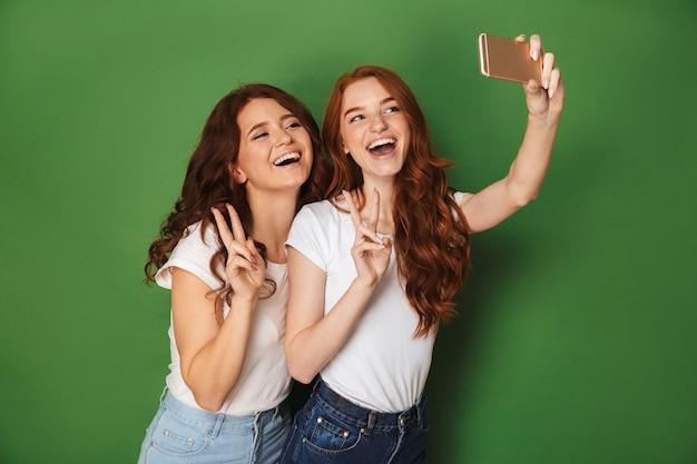 Ritratto di due donne allegre con capelli allo zenzero prendendo selfie sullo smartphone e mostrando il segno di vittoria, isolato su sfondo verde