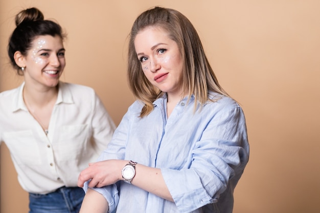 Ritratto di due donne felici in abiti bianchi e neri che sorridono alla telecamera con le braccia incrociate isolate su sfondo beige