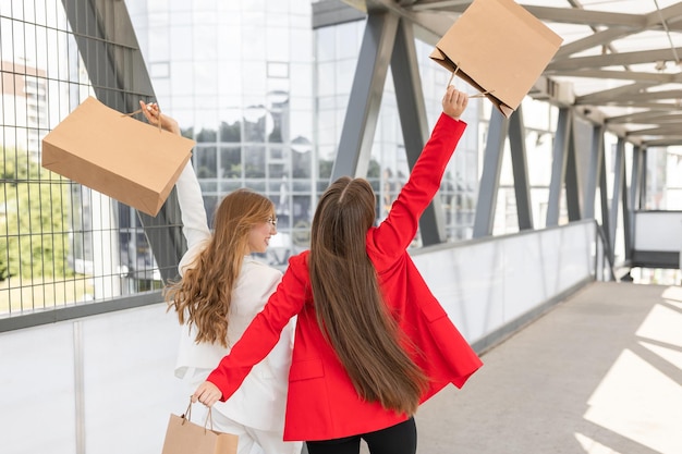 Ritratto di due donne felici vestite in abiti eleganti che camminano insieme vicino al centro commerciale con un sacco di sacchetti di carta