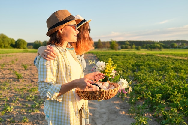 Ritratto di due donne sorridenti abbracciate madre e figlia adolescente con cesto di uova di fattoria in cappelli, con bouquet di fiori in giardino, natura estiva, sfondo tramonto, spazio copia