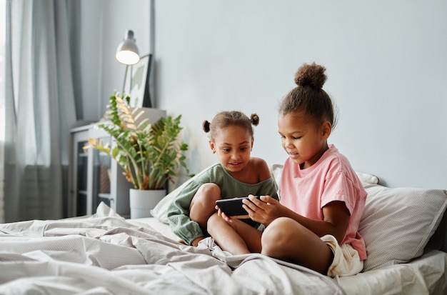 Ritratto di due ragazze afroamericane che usano lo smartphone insieme sul letto in una stanza accogliente spazio copia