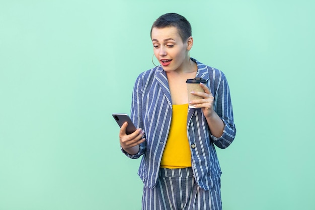 Ritratto di eccitata bella con i capelli corti giovane donna blogger in abito a righe in piedi, leggendo notizie interessanti e bevendo caffè, guardando il telefono. Interno, isolato, girato in studio, sfondo verde