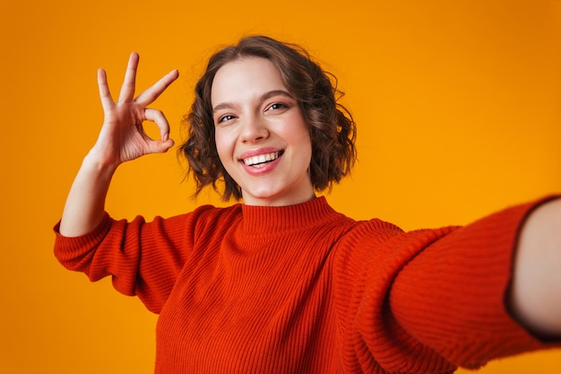 Ritratto di eccitato felice emotivo giovane donna graziosa in posa isolato su muro giallo prendere un selfie che mostra il gesto giusto.