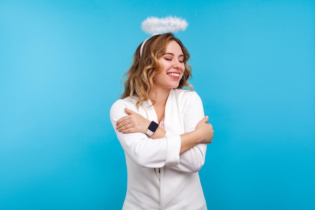 Ritratto di egoista bella donna angelica con alone sui suoi capelli ondulati e in abiti bianchi che si abbraccia, in piedi con gli occhi chiusi dal piacere e dalla felicità. girato in studio, sfondo blu