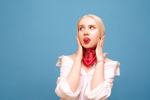 ritratto di elegante bionda in camicia bianca e velo rosso in posa davanti alla telecamera con la lingua esposta