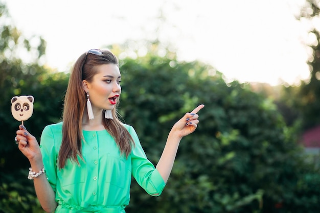 Ritratto di emotiva e bella ragazza bruna in abito verde, che tiene caramelle come un panda sul bastone e urla sorpresa. Donna graziosa ed elegante con labbra rosse, capelli lunghi che puntano a lato.