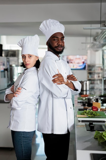 Ritratto di esperti di gastronomia in piedi schiena contro schiena all'interno della cucina professionale del ristorante mentre sorridono fiduciosi alla telecamera. Diversi lavoratori dell'industria alimentare che posano per la macchina fotografica.