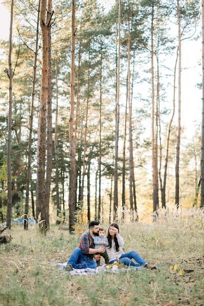 Ritratto di famiglia all'aperto di stile di vita di giovani genitori con figlio bambino, divertirsi insieme, seduti sulla coperta nella bellissima foresta di autunno al giorno pieno di sole