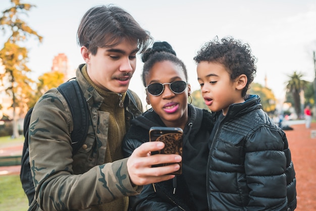 Ritratto di famiglia etnica bella razza mista divertendosi, rilassarsi e utilizzando il telefono cellulare presso il parco all'aperto.