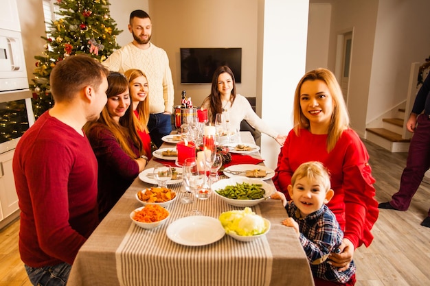 Ritratto di famiglia felice e amici seduti attorno a un tavolo festivo. Madre che lega il suo simpatico figlio sorridente, entrambi guardando la telecamera.