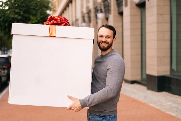 Ritratto di fattorino sorridente che tiene una grande scatola festiva con fiocco rosso in una strada cittadina all'aperto, guardando la fotocamera. Corriere maschio consegna regalo al cliente. Ragazzo che fa una sorpresa per la fidanzata o la moglie.