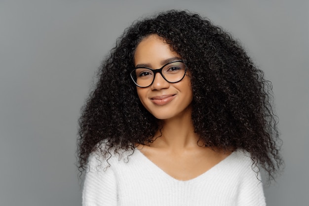 Ritratto di felice bella donna afro con folti capelli ricci, guarda attraverso gli occhiali trasparenti